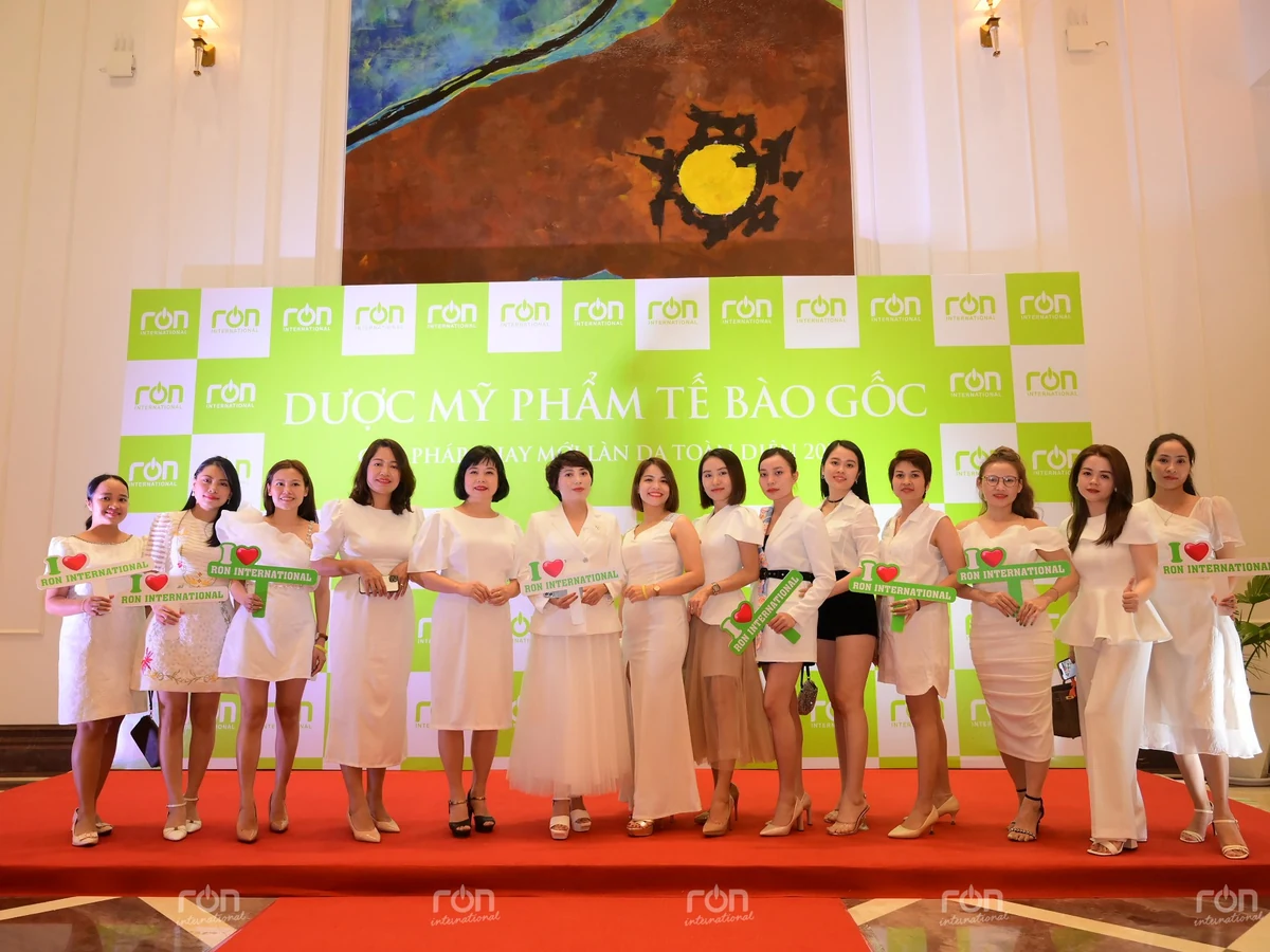 Các thành viên nữ trong trang phục trắng đứng trước phông sự kiện RON INTERNATIONAL®.