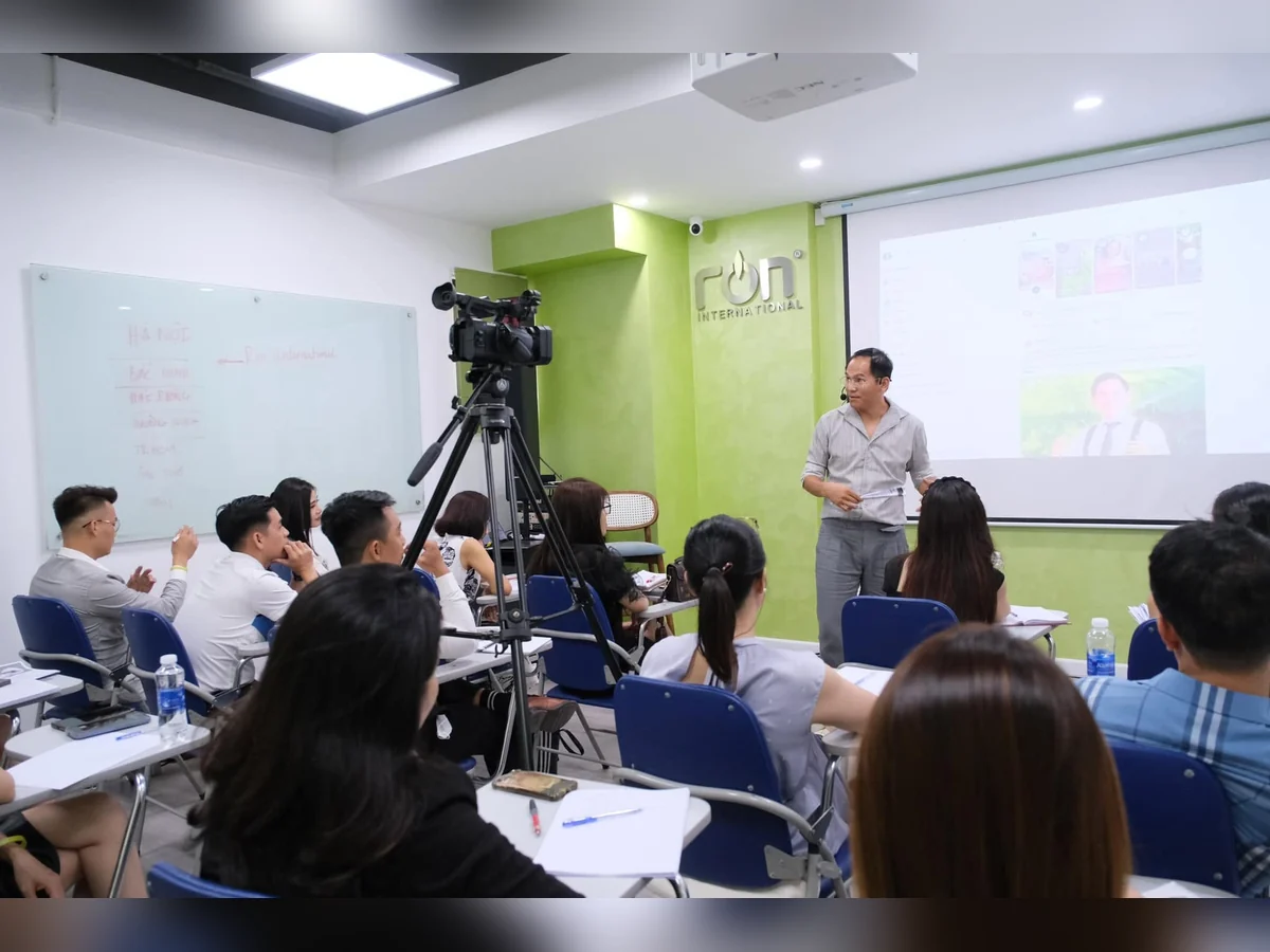 Diễn giả chia sẻ trước màn chiếu trong phòng học, học viên ngồi quanh bàn và có máy quay.