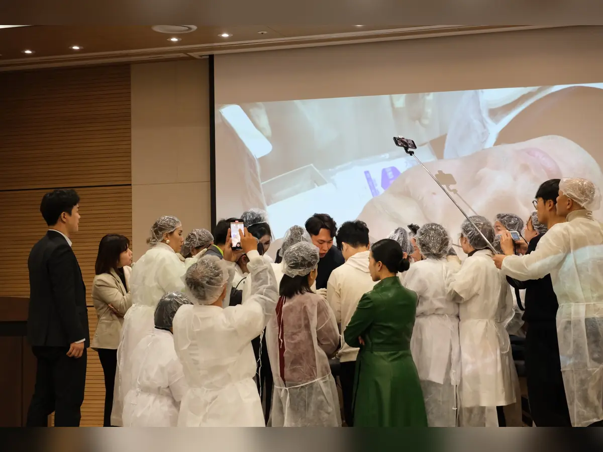 Học viên mặc trang phục bảo hộ đứng quanh bàn hướng dẫn, phía sau có màn chiếu minh họa.