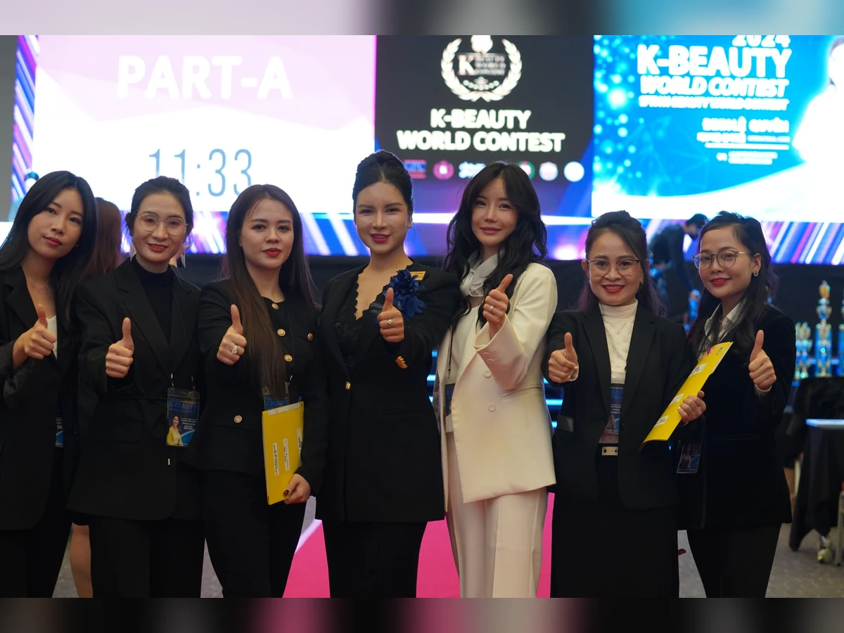 Nhóm thành viên chụp ảnh trước màn hình K-Beauty World Contest, có người cầm hoa và tài liệu.